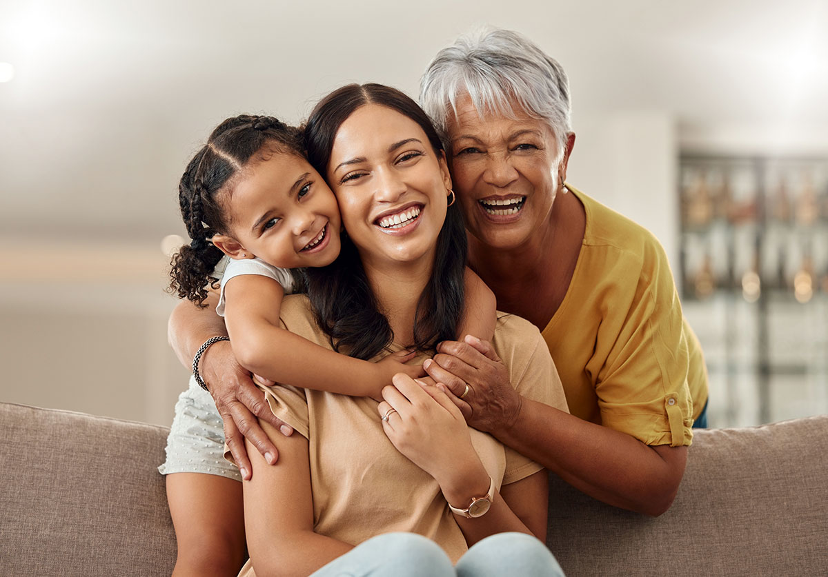 Three generations of women — a grandmother, mother, and young granddaughter — laughing together on a couch at home